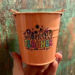 May include: A small, peach-colored metal bucket with a wire handle. The bucket features a colorful floral design and the word "TEACHER" in a playful font, with the phrase "helping little minds grow" below.