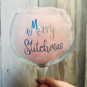 May include: A clear glass gin glass with a pink tulle bow and the text "Merry Stitchmas" in silver lettering.