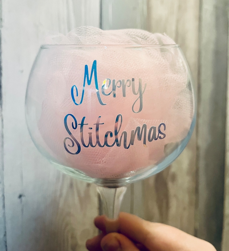 May include: A clear glass gin glass with a pink tulle bow and the text "Merry Stitchmas" in silver lettering.