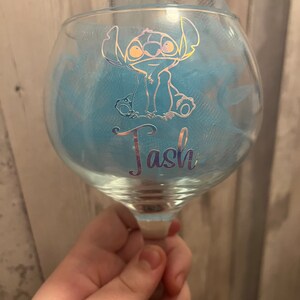 May include: A clear glass with a blue tulle bow and a gold glitter design of Stitch from Lilo & Stitch. The glass has the name "Tash" written on it.