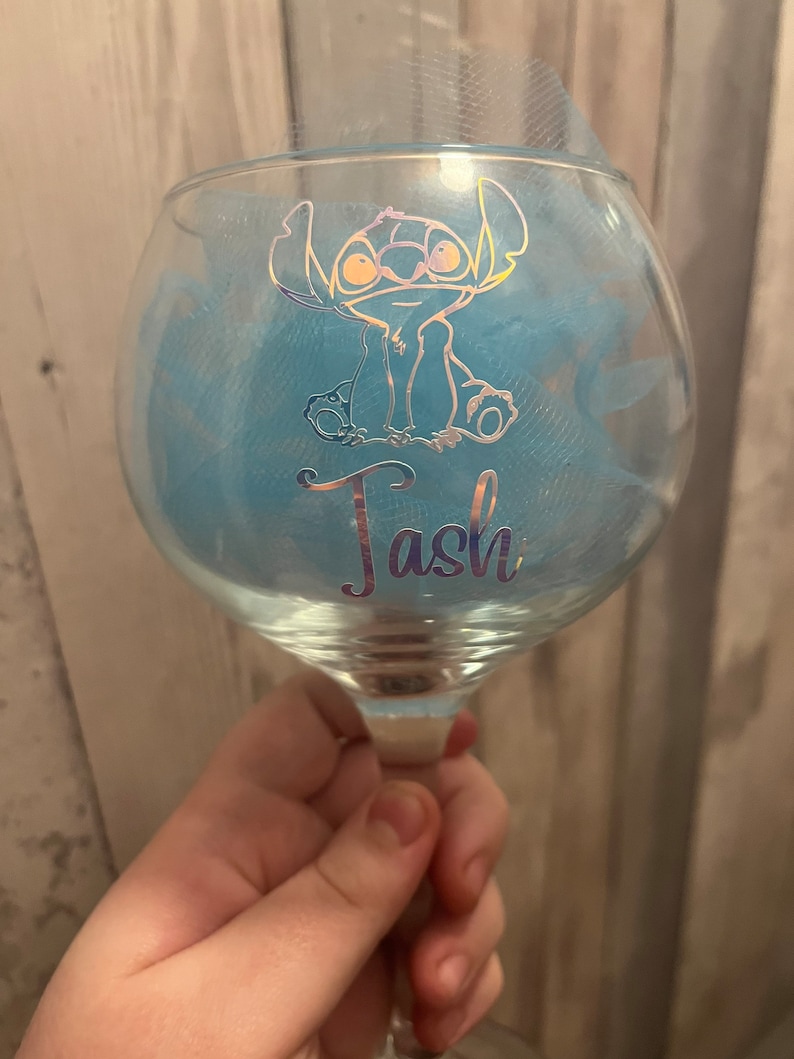 May include: A clear glass with a blue tulle bow and a gold glitter design of Stitch from Lilo & Stitch. The glass has the name "Tash" written on it.