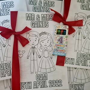May include: Four coloring books with a red ribbon and a pack of crayons. Each book features a cartoon couple with the text "Mr. & Mrs. Gaines" and the date "15th April 2022". The names "Aedi", "Cam", "Logan", and "Penny" are also included.
