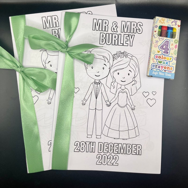 May include: Two white coloring books with a green ribbon tied around them. The front cover of the coloring book has a cartoon image of a bride and groom. The text on the front cover reads "MR & MRS BURLEY 28TH DECEMBER 2022". A small box of crayons is included.