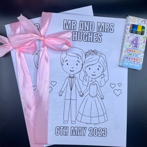 May include: A coloring book with a bride and groom illustration, a pink ribbon, and the text "MR AND MRS HUGHES 6TH MAY 2023". The book comes with a pack of four color wax crayons.