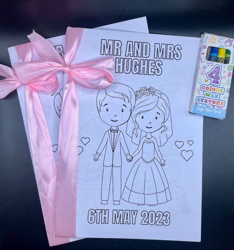 May include: A coloring book with a bride and groom illustration, a pink ribbon, and the text "MR AND MRS HUGHES 6TH MAY 2023". The book comes with a pack of four color wax crayons.