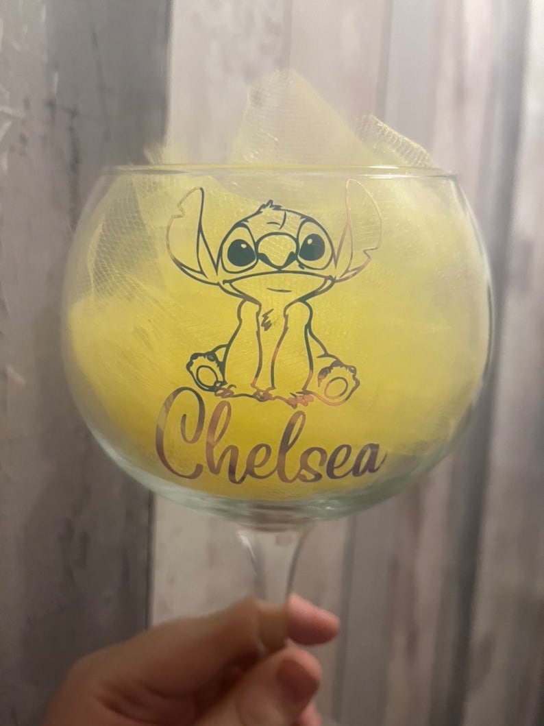 May include: A clear glass with a yellow and white design. The glass features a cartoon character with big eyes and the name "Chelsea" written on the bottom.