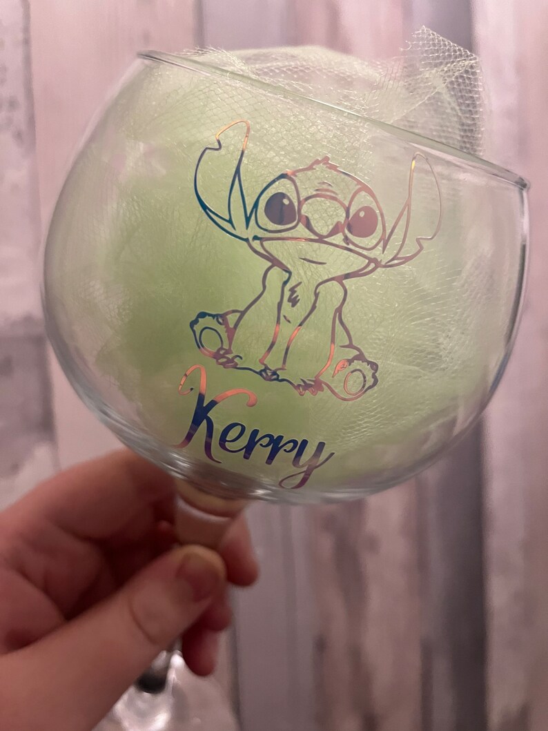 May include: A clear glass with a green and gold design of a cartoon character. The character is Stitch from the Disney movie Lilo & Stitch. The glass has the name "Kerry" written on it.