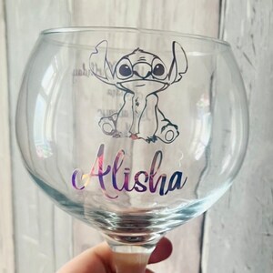 May include: A clear glass with a round bowl and a stem. The glass has a black and white cartoon character of Stitch from Lilo & Stitch on it. The name Alisha is written in a rainbow font on the glass.