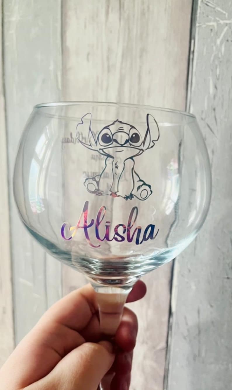 May include: A clear glass with a round bowl and a stem. The glass has a black and white cartoon character of Stitch from Lilo & Stitch on it. The name Alisha is written in a rainbow font on the glass.
