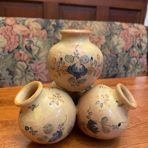 May include: Three small, round, ceramic vases with a light yellow base color. Each vase features hand-painted floral designs in blue, green, and orange. The vases are arranged in a pyramid shape, showcasing their unique, decorative style.