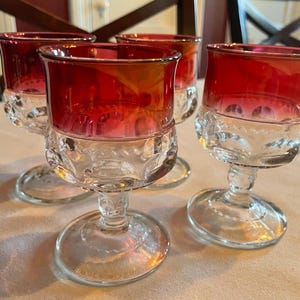 May include: Four clear glass goblets with a red-tinted upper portion. The goblets have a decorative pattern around the middle and a round base. The glasses are arranged on a light-colored surface.