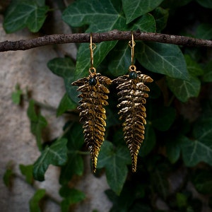 May include: Gold-coloured fern leaf earrings with a small dark green stone. The earrings hang from a thin, brown branch, set against a backdrop of green ivy.
