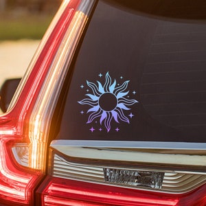 Peut inclure: Une voiture blanche avec un autocollant de soleil bleu et violet sur la vitre arrière. L'autocollant représente un soleil avec des rayons et des étoiles autour.