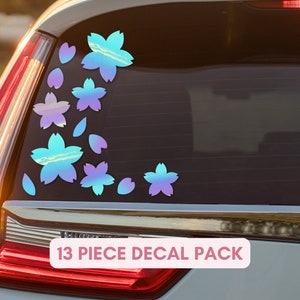 May include: A set of 13 iridescent flower and petal decals applied to a car window. The decals shift colors from blue to purple. The text "13 PIECE DECAL PACK" is displayed in a pink box.