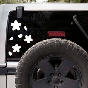 May include: White cherry blossom decals on a vehicle's rear window. The decals include three flowers and several falling petals. The vehicle is silver with a black window and a spare tire mounted on the back.