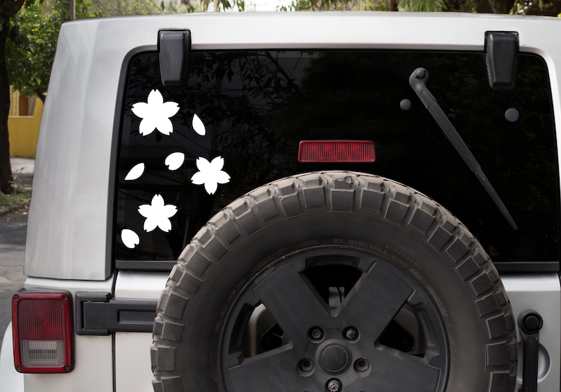 May include: White cherry blossom decals on a vehicle's rear window. The decals include three flowers and several falling petals. The vehicle is silver with a black window and a spare tire mounted on the back.