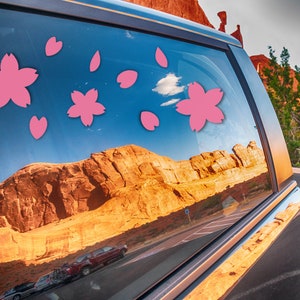 May include: A car window adorned with pink cherry blossom stickers. The window reflects a scenic desert landscape with red rock formations and a blue sky. The stickers add a touch of floral charm to the vehicle.