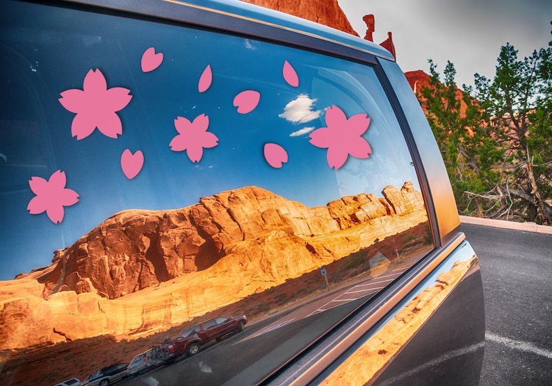 May include: A car window adorned with pink cherry blossom stickers. The window reflects a scenic desert landscape with red rock formations and a blue sky. The stickers add a touch of floral charm to the vehicle.