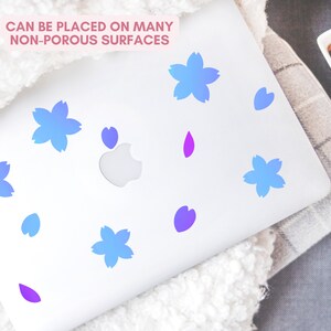 May include: A white laptop adorned with blue and purple flower and petal-shaped decals. The text "CAN BE PLACED ON MANY NON-POROUS SURFACES" is displayed in pink. The decals are arranged across the laptop's surface.