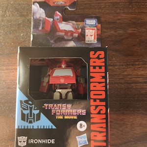 May include: A red and white Ironhide Transformers action figure in its original packaging. The box features the Studio Series logo, the text "TRANSFORMERS THE MOVIE", and the Hasbro logo. The packaging is black, blue, and red.