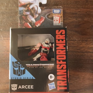 May include: A boxed Arcee action figure from the Transformers Studio Series. The figure is red, white, and gray. The box is black with red and blue accents and the text "Transformers" and "Arcee". The packaging also includes the text "Rise of the Beasts".