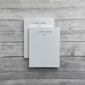 May include: Two white note cards with the text "A Love Note for you" printed on them. The cards are stacked on top of each other and are sitting on a light wood surface.