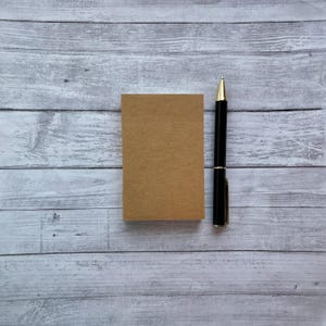 May include: A rectangular, blank, light brown kraft paper notepad positioned vertically next to a sleek black ballpoint pen with a gold-colored tip, clip, and center band, featuring a smooth, matte finish on the paper surface.