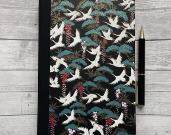 May include: Black journal with a cover featuring a repeating pattern of white cranes flying over stylized green trees and gold accents. The journal has a black spine and a red bookmark.