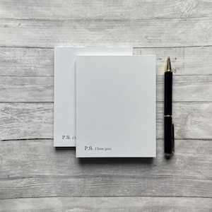 May include: Two blank white note cards with the text "P.S. I love you." printed on the bottom card. A black pen is lying on a wooden surface next to the cards.