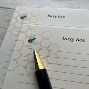 May include: Two white notepads with a honeycomb pattern and a bee illustration. The text "busy bee" is printed on the top right corner of each notepad. A black and gold pen is placed on the notepads.