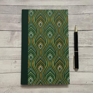 May include: A hardcover notebook with a green and gold patterned cover and a dark green spine. A black pen with gold accents rests beside the notebook on a light wood surface. The notebook's design features a repeating, stylized floral pattern.