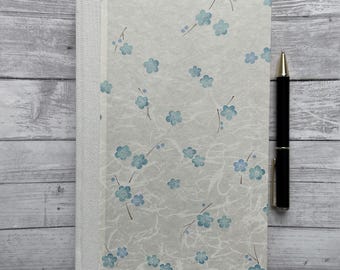 May include: A white journal with a blue floral pattern and a white spine. The journal is lying on a light gray wooden surface with a black pen next to it.