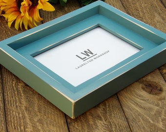 May include: A teal blue picture frame with a white mat and a white label with the text "LW LAURELTON WORKSHOP".