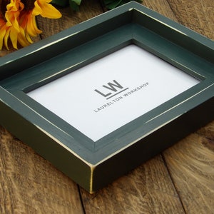 May include: A dark green picture frame with a white border and the text "LW LAURELTON WORKSHOP" on the inside.