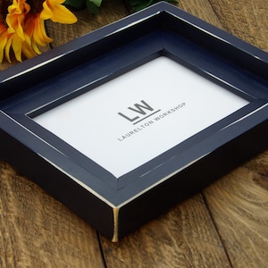 May include: A navy blue picture frame with a white distressed finish. The frame has a white mat and the text "LW LAURELTON WORKSHOP" is printed on the mat.