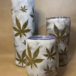 May include: A set of three insulated tumblers with a white and gray marble effect. Each tumbler features a repeating pattern of gold-colored cannabis leaves. The set includes a tall, cylindrical tumbler, a shorter tumbler, and a stemless wine glass.