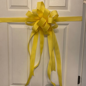 May include: A large yellow bow tied on a white door. The bow is made of satin ribbon and has long tails.