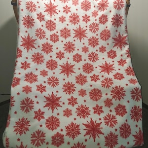 May include: A white blanket featuring a repeating pattern of red snowflakes. The snowflakes vary in size and design, creating a festive winter theme. The blanket is draped over a wooden chair.
