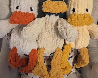 Puede incluir: Tres peluches de pato hechos a mano con ganchillo en blanco, verde y amarillo. Cada pato tiene un pico y patas de color naranja o amarillo contrastantes, con ojos negros. Los patos están colocados sobre una tela estampada, creando una estética artesanal y acogedora.