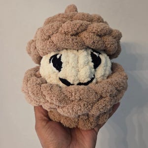 May include: A plush, crocheted acorn toy featuring a textured, light brown cap and a cream-colored base. The acorn has embroidered black eyes and a black mouth, creating a simple facial expression. The yarn is thick, soft, and fuzzy in appearance.