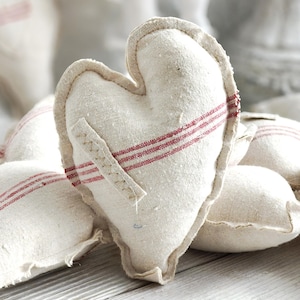 Può includere: Primo piano di diversi cuscini di stoffa a forma di cuore, bianco sporco, con strisce rosse. I cuscini hanno un bordo cucito e una piccola toppa di stoffa. Lo sfondo è sfocato, con altri cuscini e una statua bianca visibili.