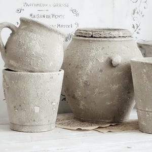 May include: A collection of weathered, gray ceramic pots and vessels. The assortment includes a pitcher, a large lidded pot, and several smaller containers. The pieces have a rustic, aged appearance, suggesting they are vintage or antique. The background features faded French text.