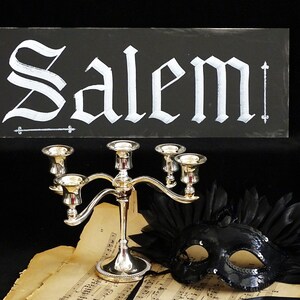 May include: A black chalkboard sign with the word "Salem" written in white calligraphy. Below the sign is a silver candelabra with five candle holders, a black masquerade mask with sequins, and a sheet of music.