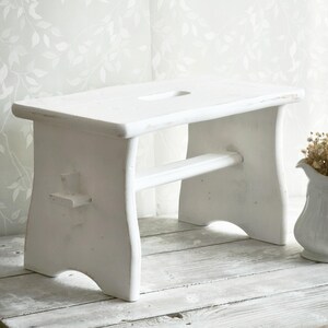 May include: A white wooden stool with a rectangular seat and a cutout. The stool has four legs and a horizontal support beam. The wood has a distressed finish. The stool is on a wooden surface. A white vase with dried flowers is to the right.