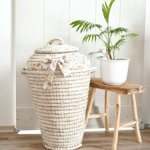May include: A large, off-white woven laundry basket with a matching lid and a floral ribbon detail. A small wooden stool holds a potted green plant. The basket has a handle and is set against a white paneled wall.