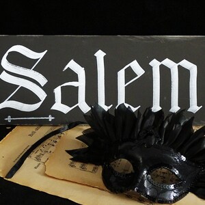 May include: A black sign with white lettering that reads "Salem" in a gothic font. A black masquerade mask with feathers is in the foreground.