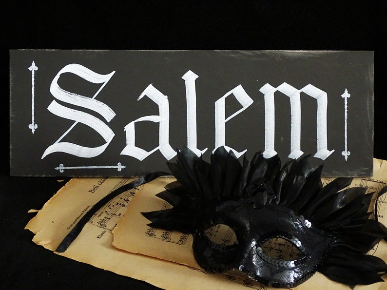 May include: A black sign with white lettering that reads "Salem" in a gothic font. A black masquerade mask with feathers is in the foreground.
