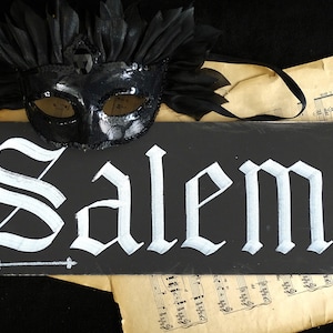 May include: A black chalkboard sign with the word "Salem" written in white calligraphy. A black masquerade mask with sequins is on top of the sign.