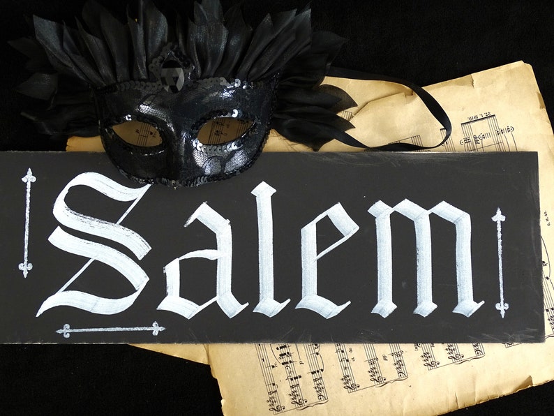 May include: A black chalkboard sign with the word "Salem" written in white calligraphy. A black masquerade mask with sequins is on top of the sign.