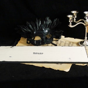 May include: A white rectangular sign with black edges. The sign has the text "Blackstone Gifts" printed on it. The sign is sitting on top of a piece of sheet music with a black masquerade mask with feathers on top of the sheet music.